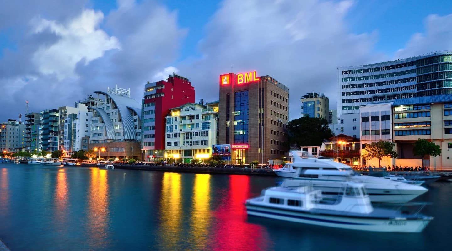 The illuminated BML sign on Bank of Maldives’ head office beside the Malé waterfront at dusk, with boats in the foreground.
