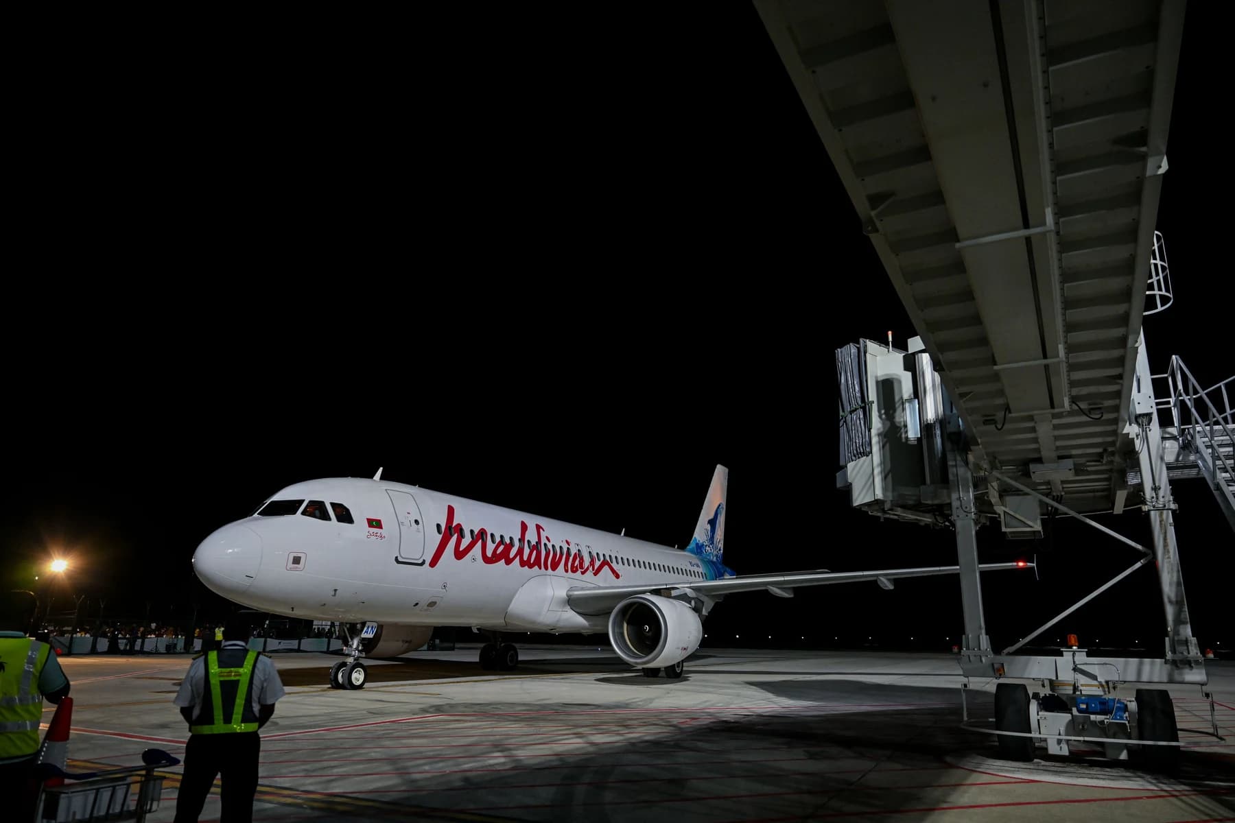 A white Maldivian Airbus A320 with red script lettering stands beside a passenger boarding bridge at night as ground staff in reflective vests watch.