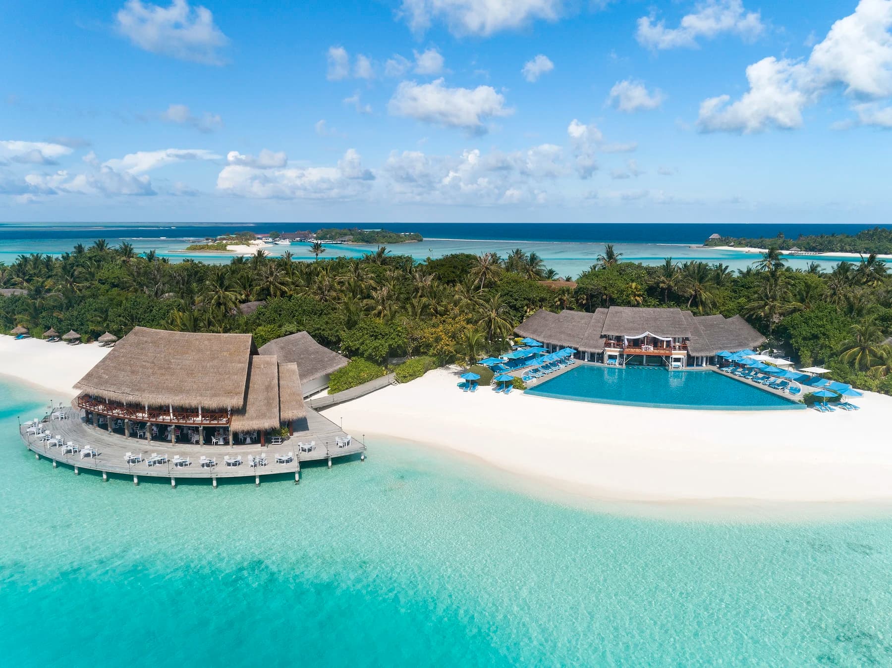 Aerial view of a palm-covered resort island with a thatched overwater restaurant, an infinity pool and a white-sand beach beside a turquoise lagoon.