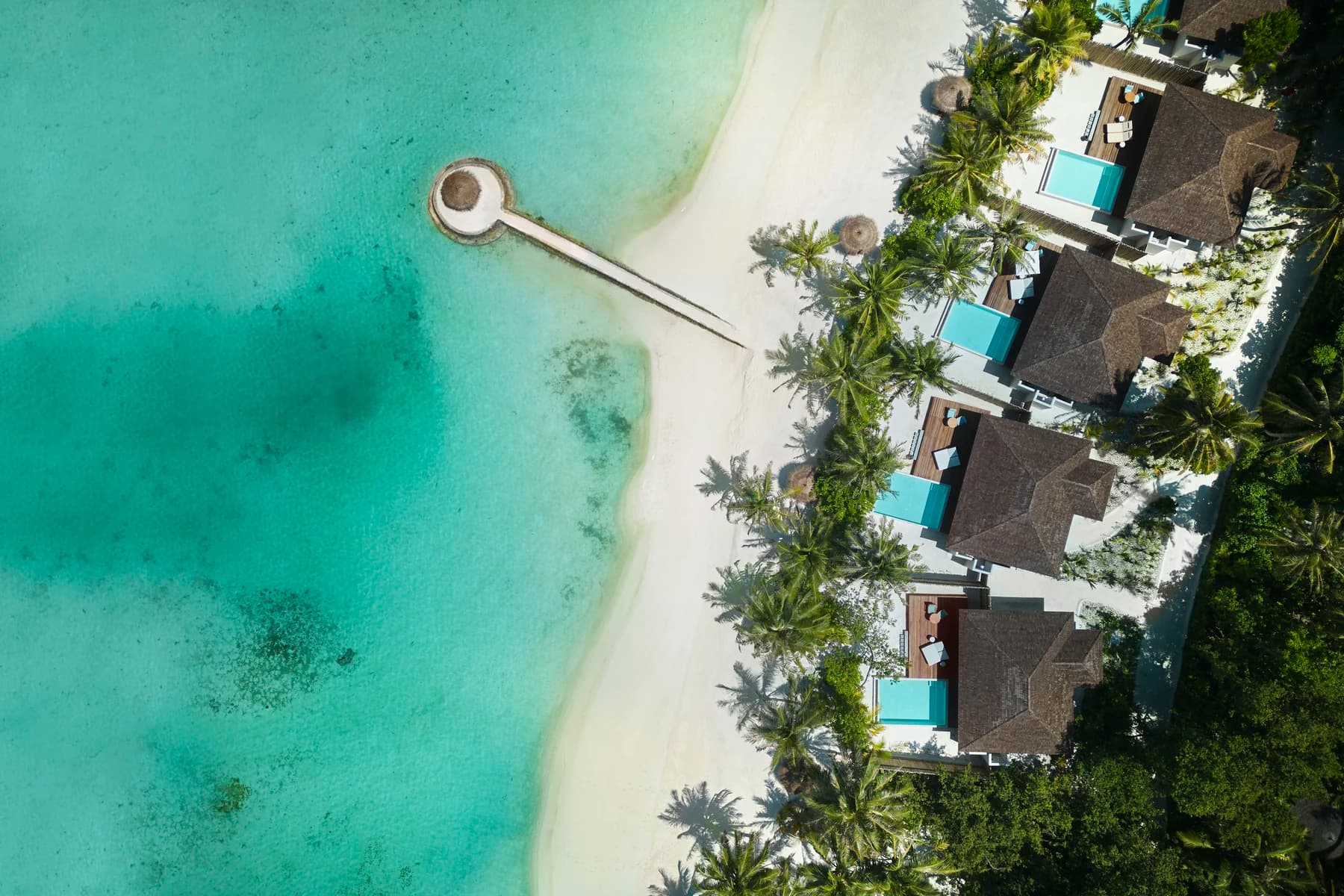 Aerial view of thatched beach villas with private pools beside a white-sand beach, a jetty and a turquoise lagoon.