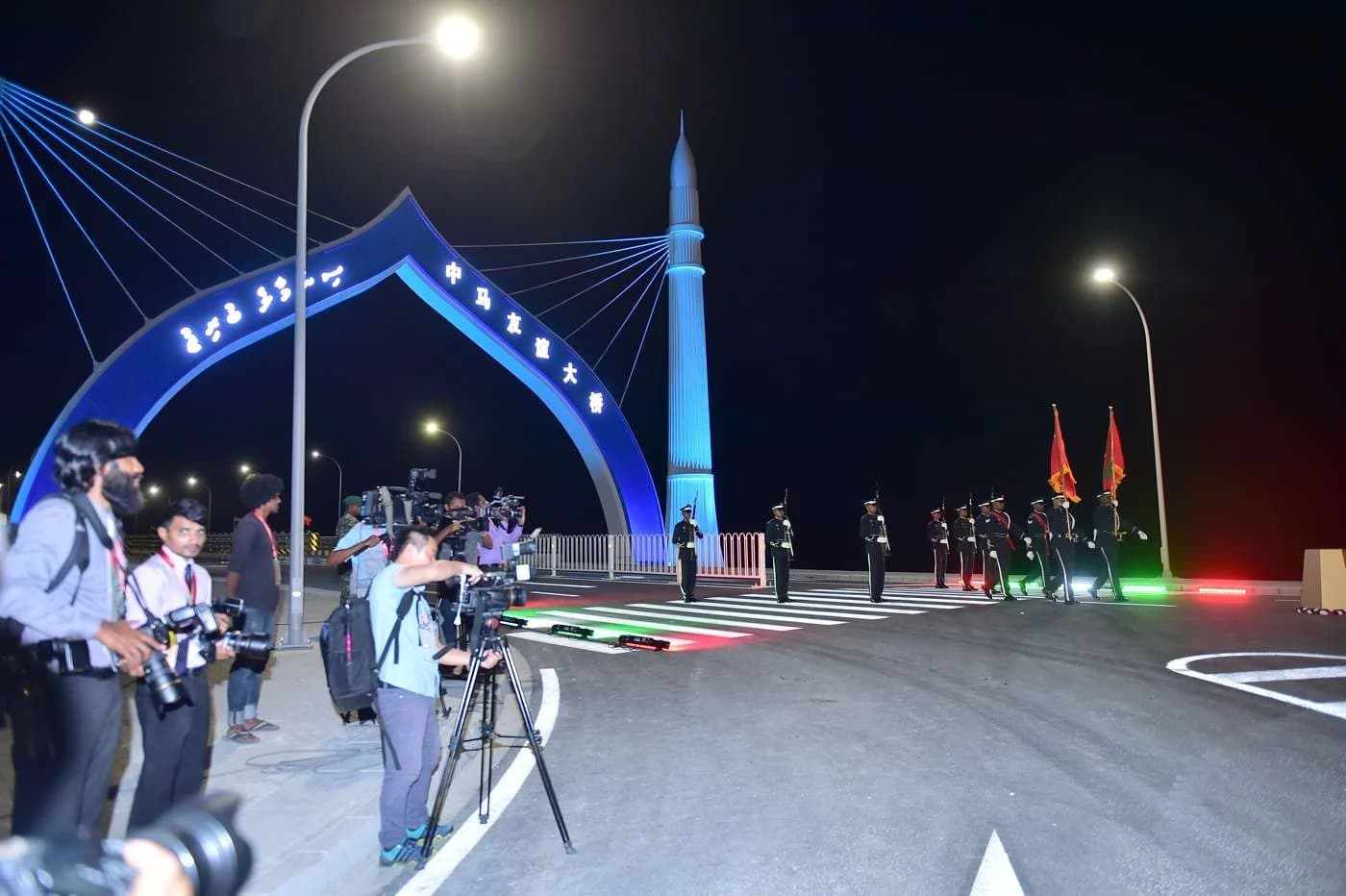 An illuminated blue arch and pylon at the entrance to a bridge at night, with photographers and an honour guard on the road.