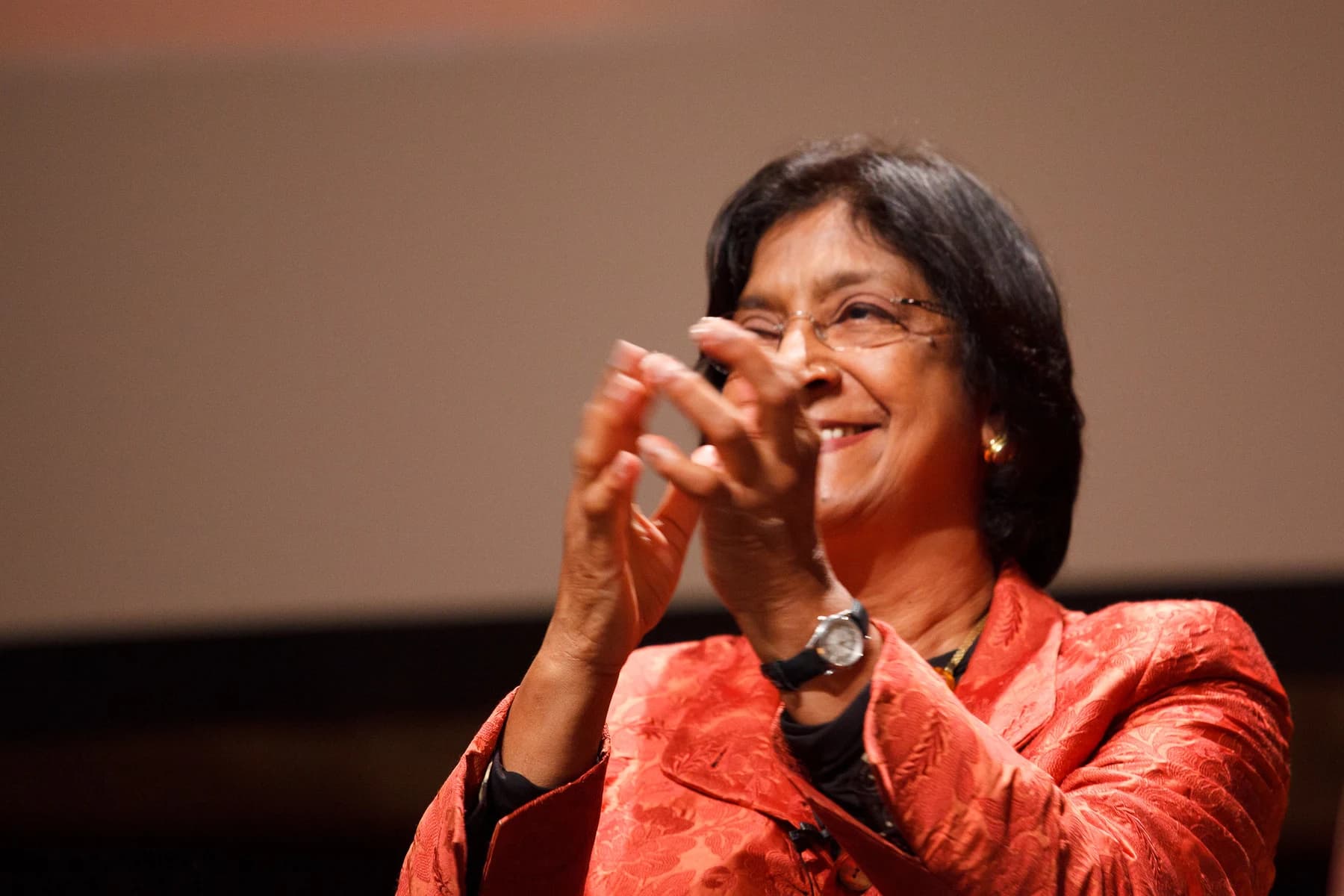 Navi Pillay, wearing glasses and a red patterned jacket, smiles and applauds on a stage