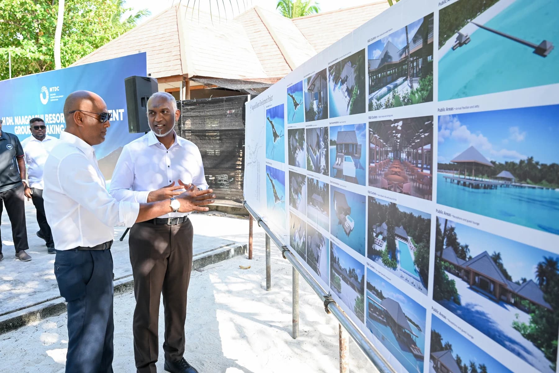 Two men in white shirts talk beside a long board of resort design images on a sandy island, with an MTDC groundbreaking banner behind them.