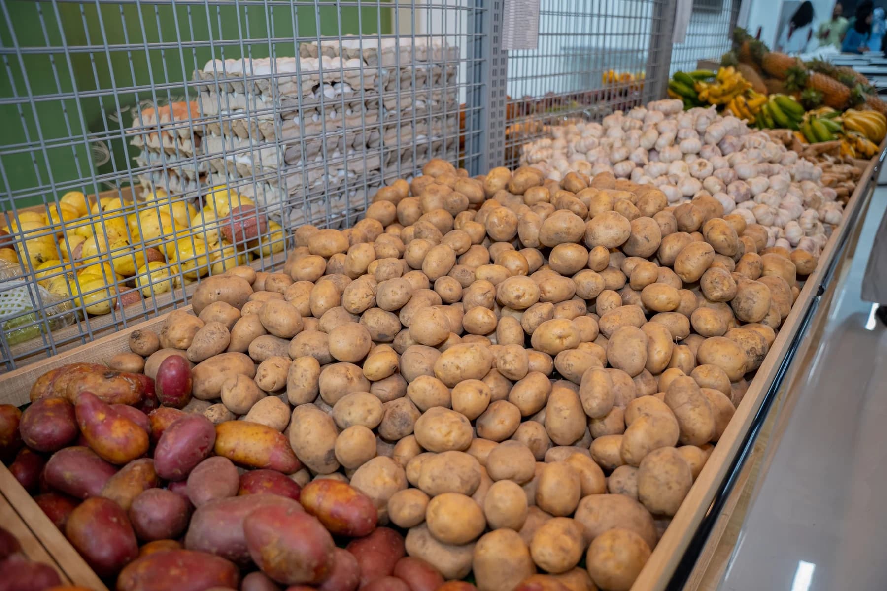 A display bin piled with potatoes and sweet potatoes, with garlic, trays of eggs, lemons, bananas and pineapples behind it in a shop.