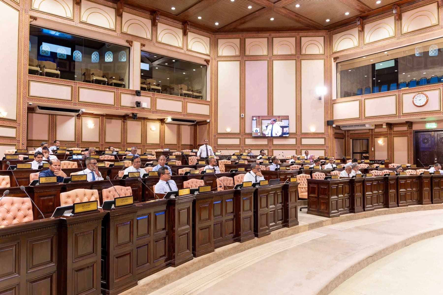 The chamber of the People's Majlis during a sitting, with members seated at wooden desks and one member standing to speak.