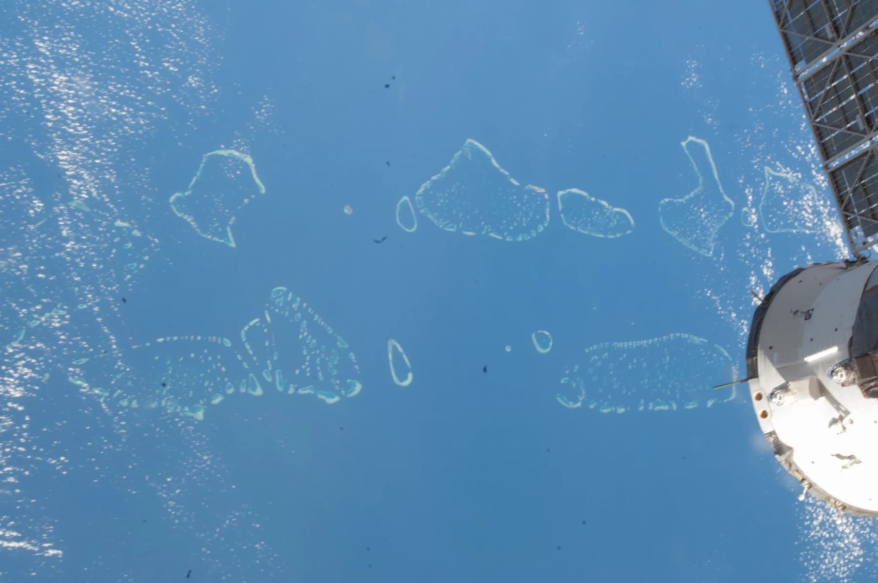 Ring-shaped coral atolls in a blue ocean seen from orbit, with the white hull of a spacecraft and a solar array at the right edge.