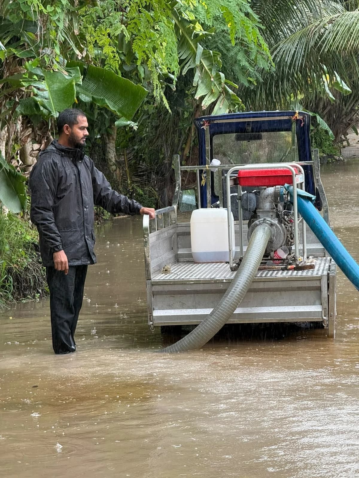 A man in a rain jacket stands in floodwater beside a water pump mounted on a small truck, with a hose running into the water.