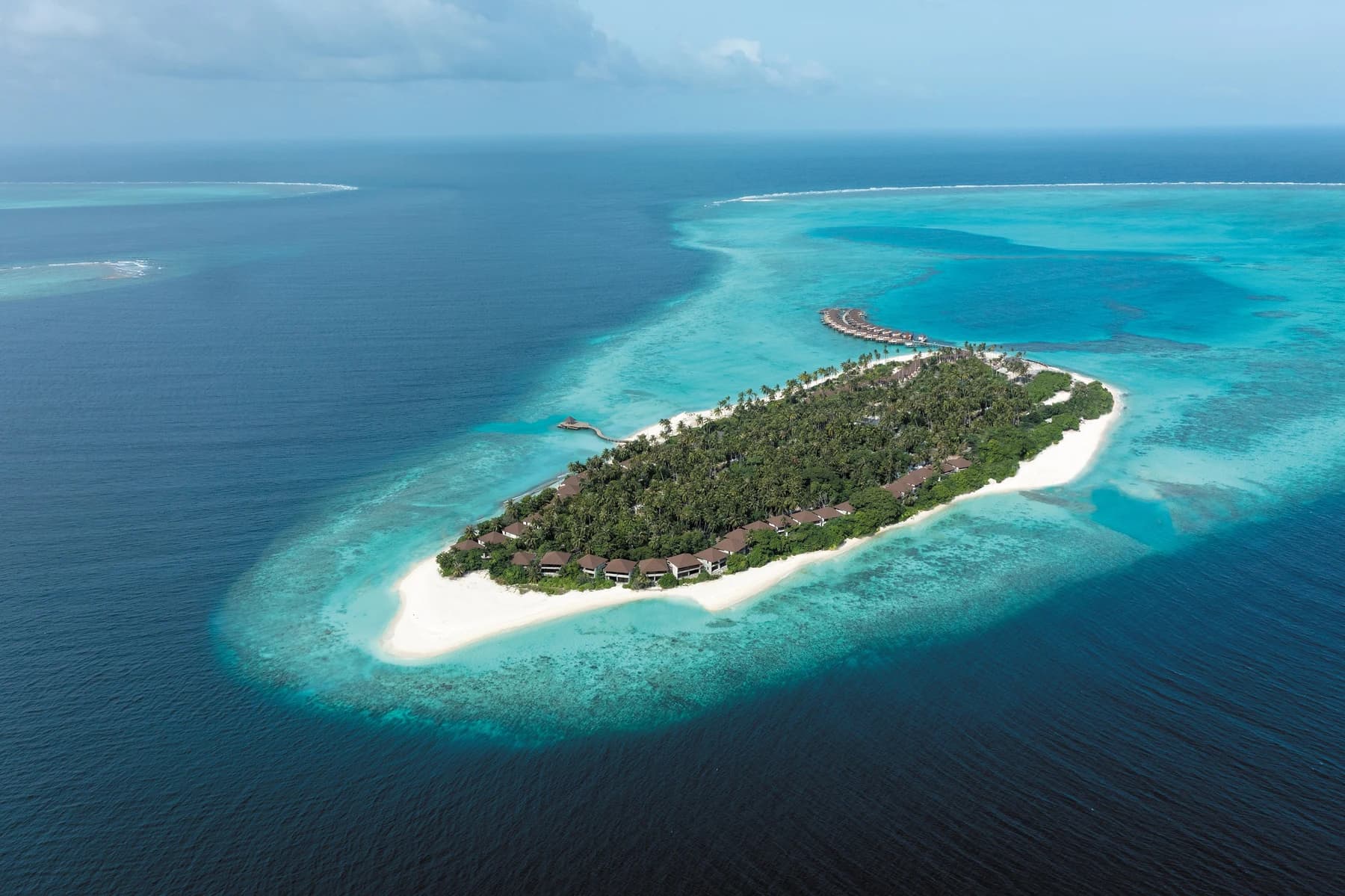 Aerial view of a palm-covered resort island ringed by white sand and a turquoise lagoon, with a row of overwater villas at one end.