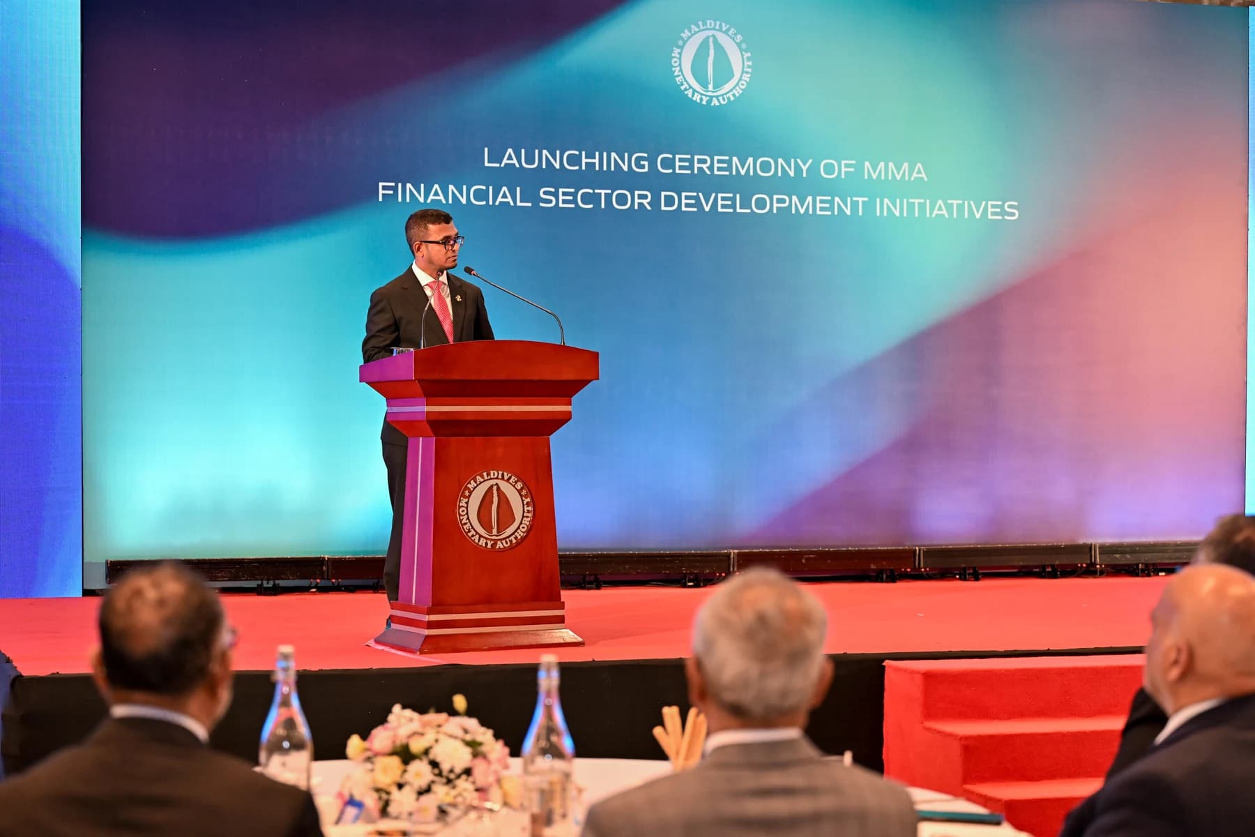 A speaker at a red Maldives Monetary Authority lectern on a stage beneath a screen reading “Launching Ceremony of MMA Financial Sector Development Initiatives”, with guests seated in the foreground.