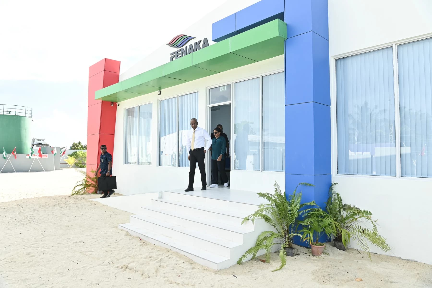 A white single-storey building with red, green and blue panels and a FENAKA sign, with people walking out of its door onto sandy ground.