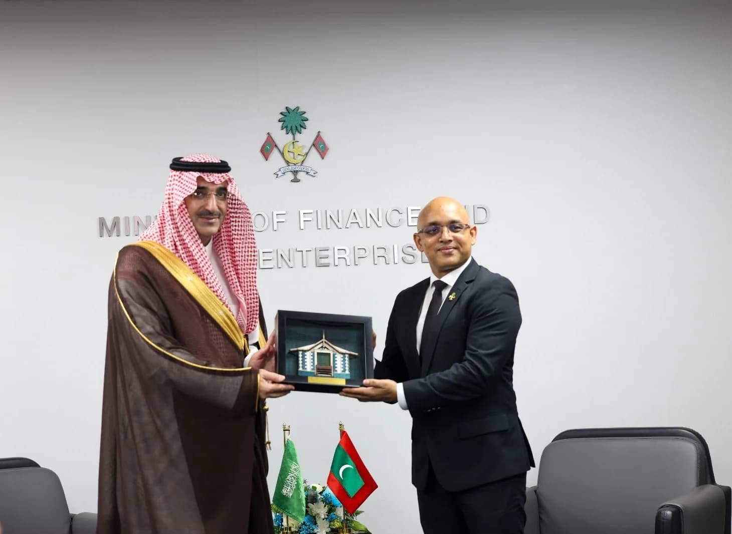 Two men stand in front of a wall bearing the Maldivian state emblem and the words Ministry of Finance and Public Enterprises, holding a framed model between them beside Saudi and Maldivian flags.