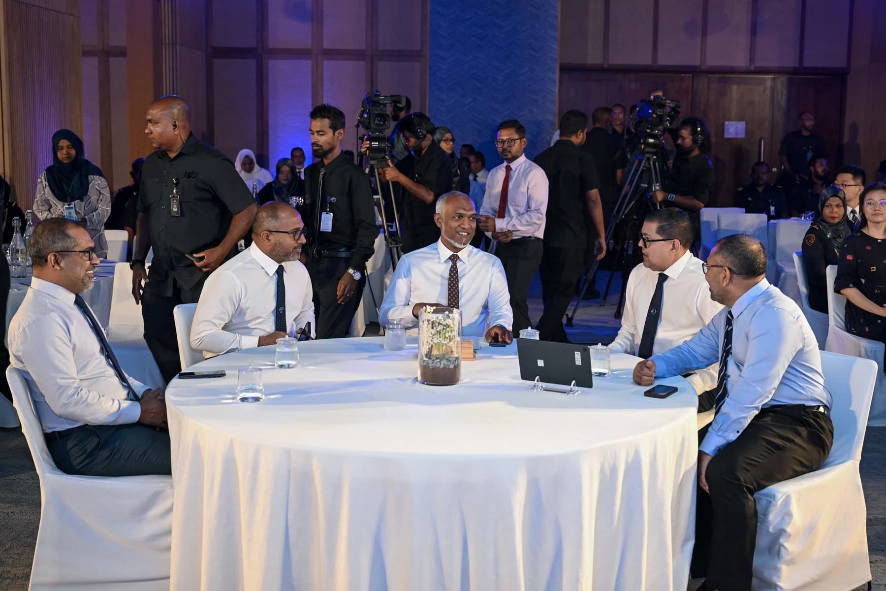 President Muizzu and four officials seated at a round table at a launch event, with camera operators and guests behind.
