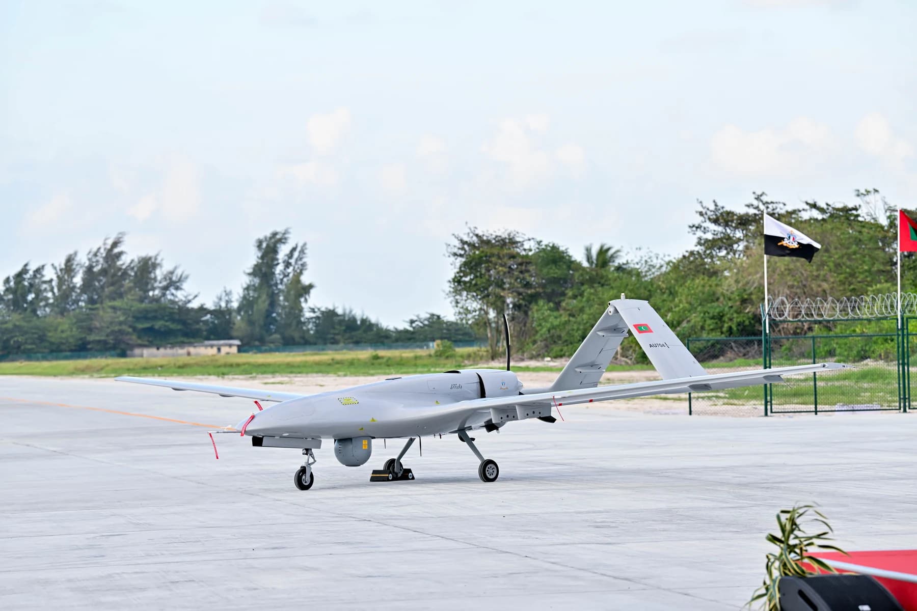 A grey military drone with a Maldivian flag on its tail parked on a runway apron, with trees and flags behind.