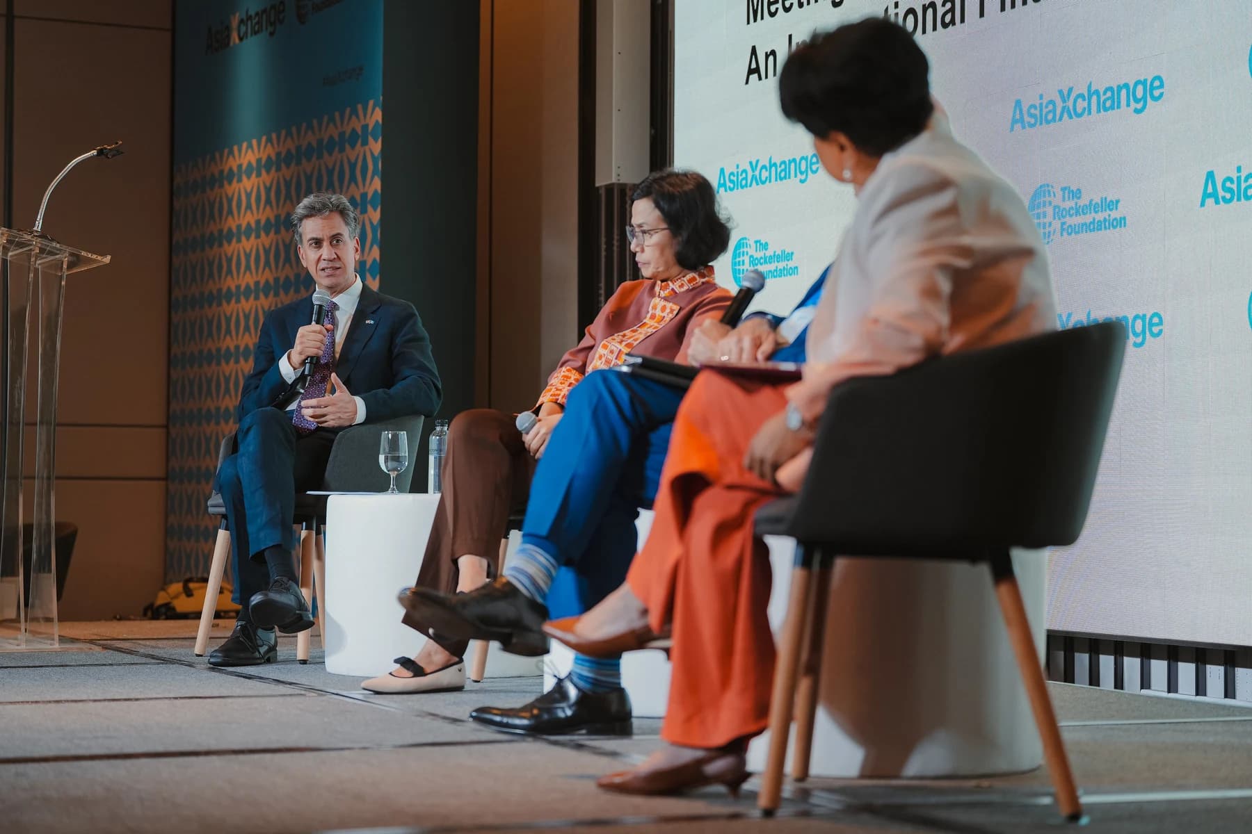 Ed Miliband, in a dark suit, speaks into a microphone on a panel stage beside other speakers, with Rockefeller Foundation and AsiaXchange signs behind