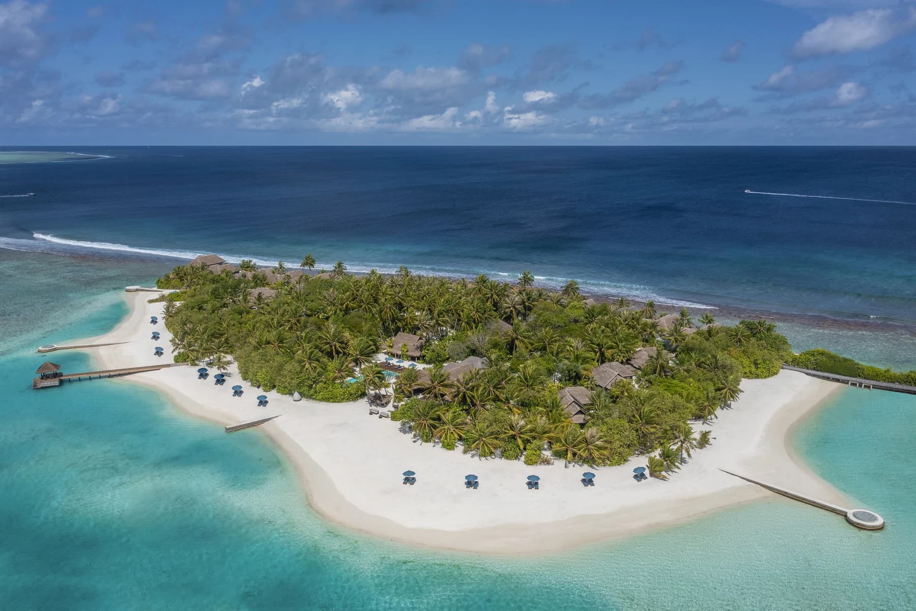 Aerial view of a small island resort ringed by white sand and turquoise lagoon, with thatched villas among palm trees and jetties reaching into the water.
