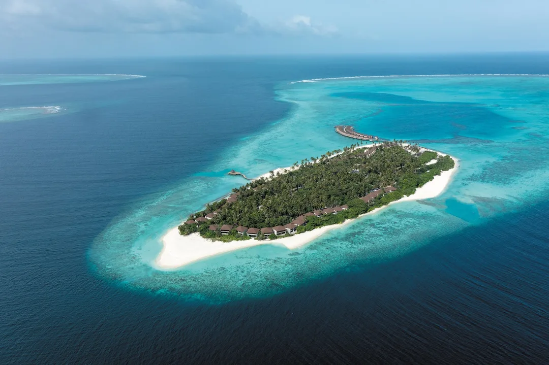 Aerial view of a palm-covered resort island ringed by white sand and a turquoise lagoon, with a row of overwater villas at one end.