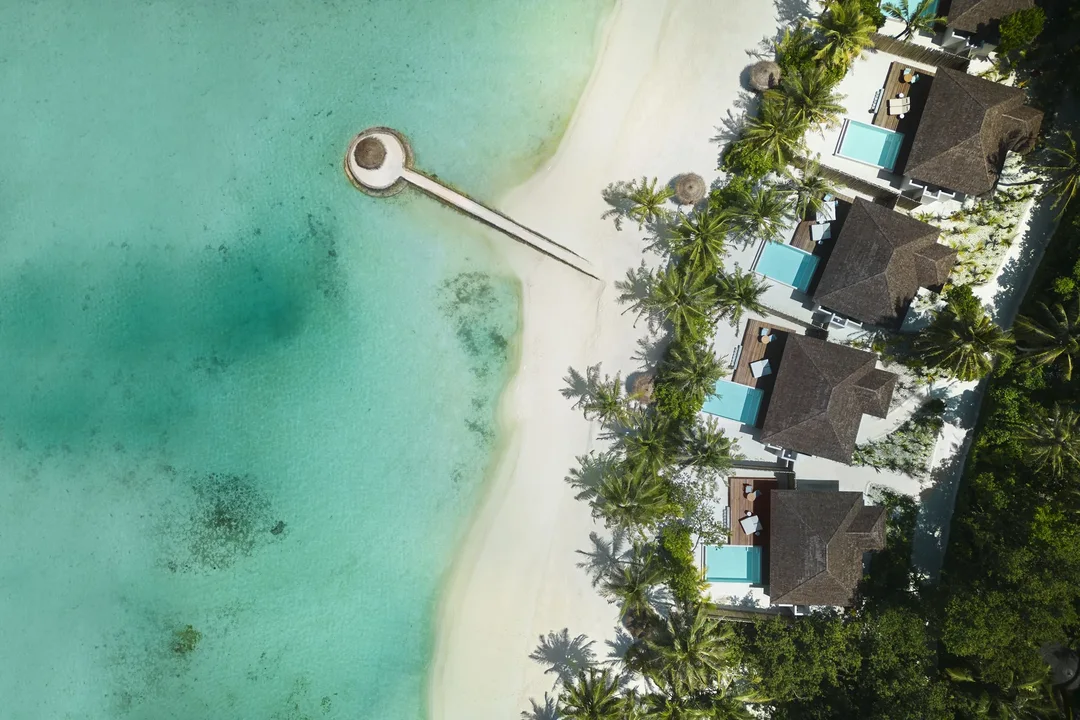 Aerial view of thatched beach villas with private pools beside a white-sand beach, a jetty and a turquoise lagoon.