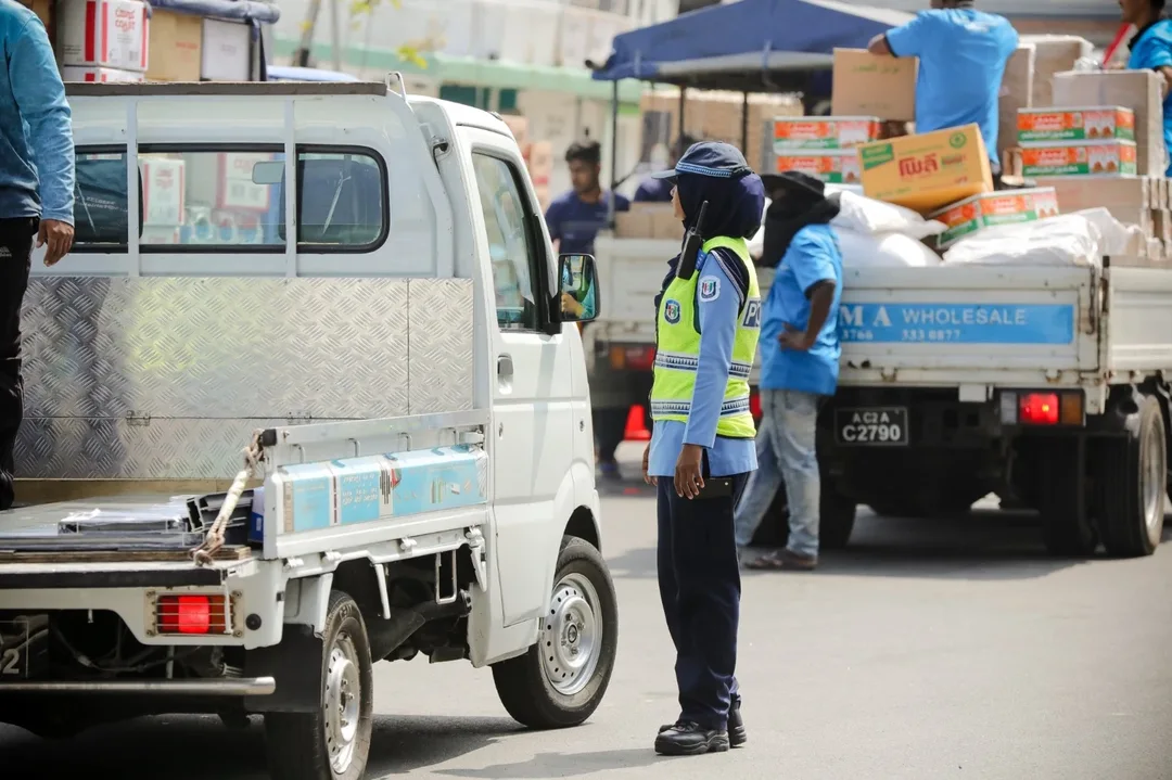 A police officer in a high-visibility vest stands on a Malé street between two small pickup lorries, one loaded with boxes and sacks by workers.