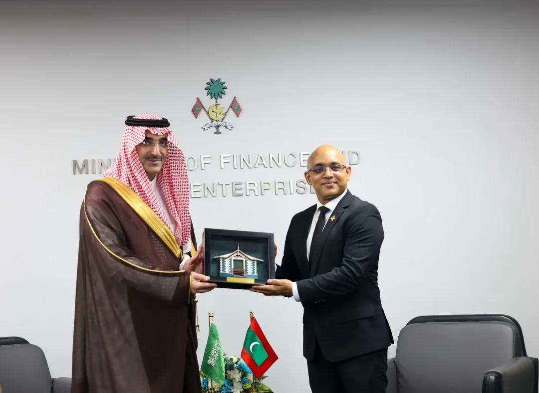 Two men stand in front of a wall bearing the Maldivian state emblem and the words Ministry of Finance and Public Enterprises, holding a framed model between them beside Saudi and Maldivian flags.