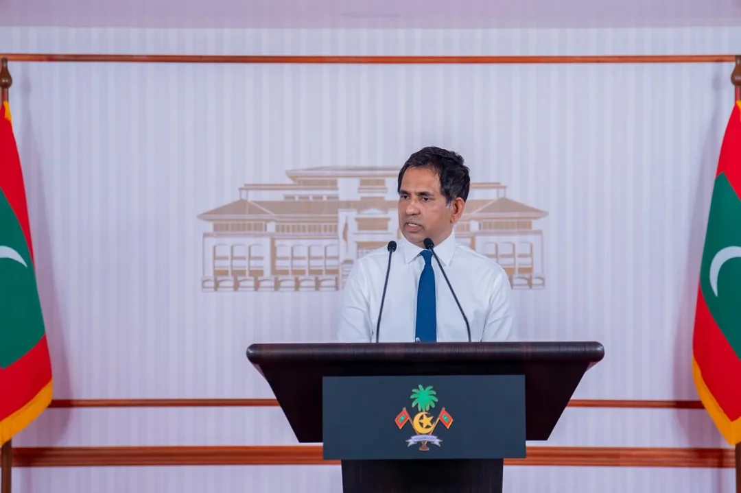 A man in a white shirt and blue tie speaks at a lectern bearing the Maldivian state emblem, between two Maldivian flags.