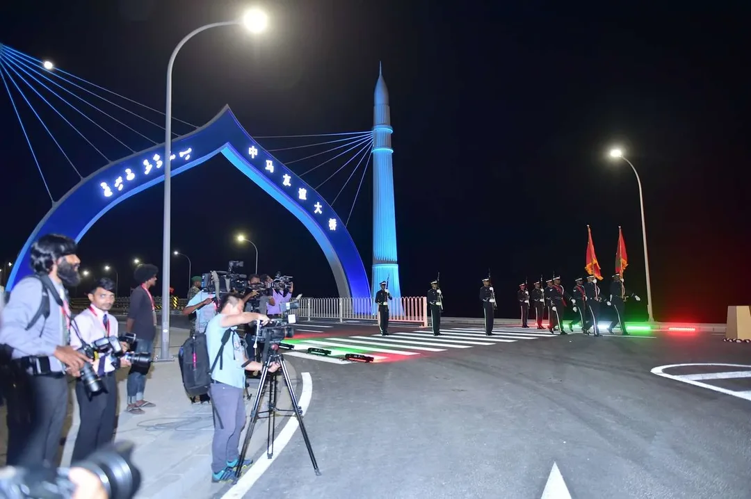 An illuminated blue arch and pylon at the entrance to a bridge at night, with photographers and an honour guard on the road.