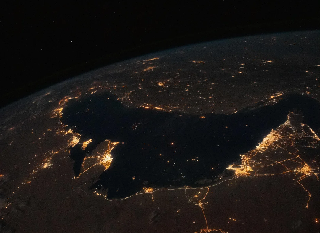 Night view from space of the Persian Gulf, a dark sea ringed by orange city lights, with scattered ship lights on the water and the Strait of Hormuz at right