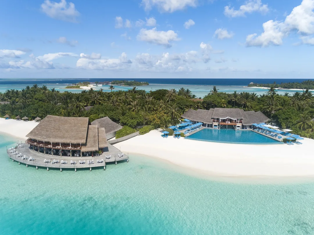 Aerial view of a palm-covered resort island with a thatched overwater restaurant, an infinity pool and a white-sand beach beside a turquoise lagoon.