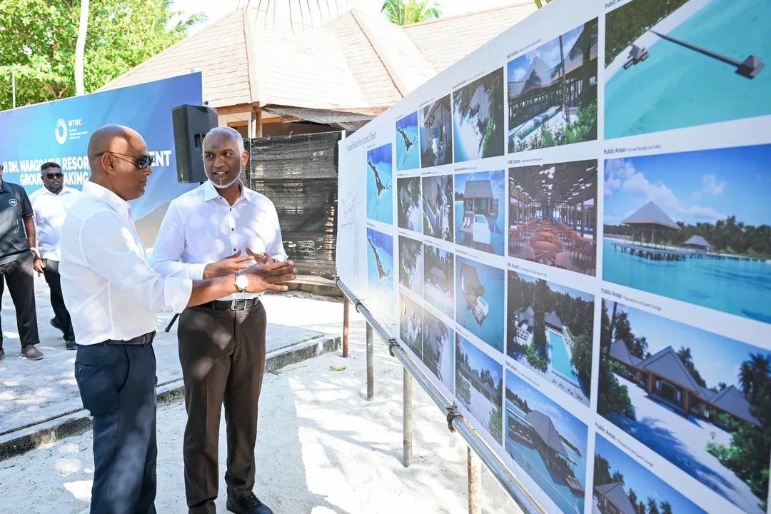 Two men in white shirts talk beside a long board of resort design images on a sandy island, with an MTDC groundbreaking banner behind them.