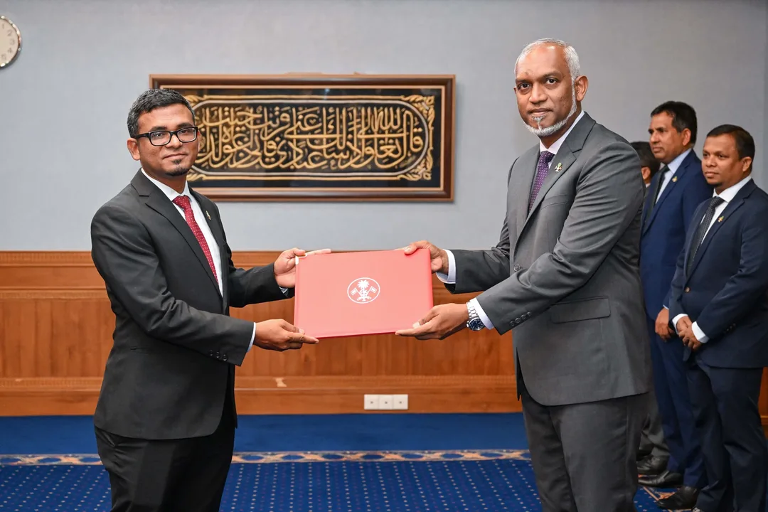 Mohamed Muizzu, in a grey suit, hands a red folder bearing the state emblem to Ahmed Munawar, in a dark suit and glasses, with officials standing behind.