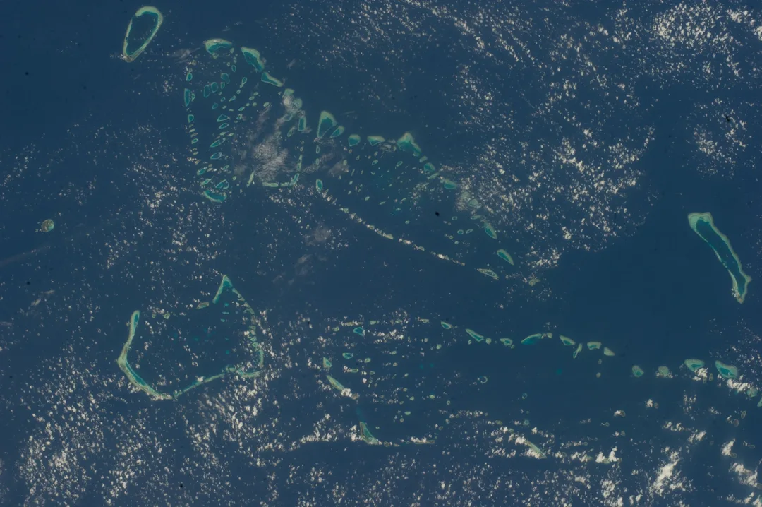 Atolls of the Maldives seen from the International Space Station, their reefs showing as pale turquoise rings and patches in dark blue ocean under scattered cloud.