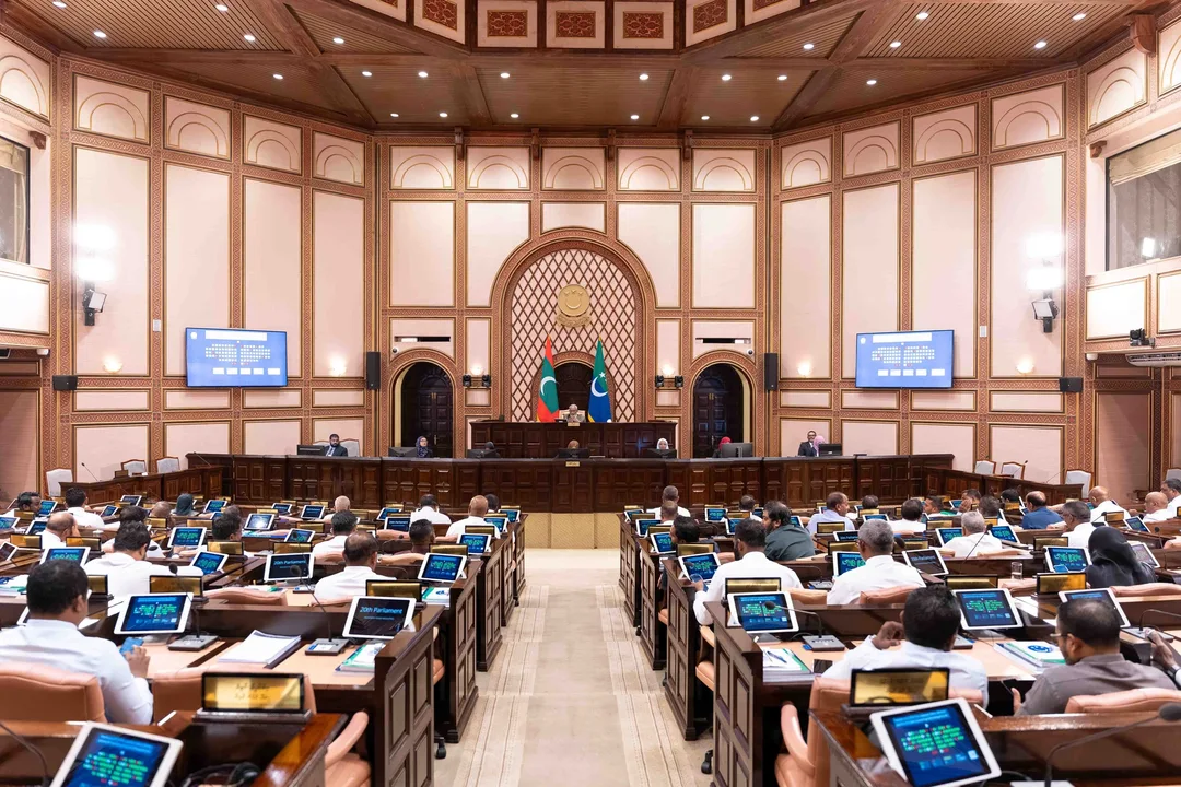 A wide view of the People's Majlis chamber from the back, with members seated at their desks facing the presiding officer's dais, which is flanked by two flags.