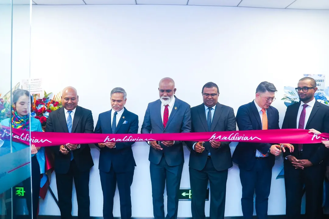 Men in suits cut a pink ribbon printed with the Maldivian logo in a white-walled office, with flower stands on either side.