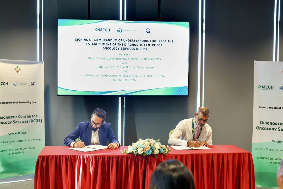 Two men sign documents at a red-draped table beneath a screen announcing the signing of an MoU for the Diagnostic Centre for Oncology Services, with MCGH, N-Vascular and Quantum Heights logos.