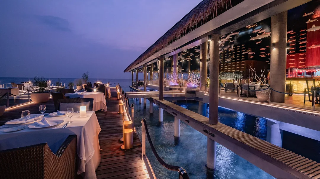 Candlelit tables with white cloths on an overwater restaurant deck at dusk, under a thatched roof.