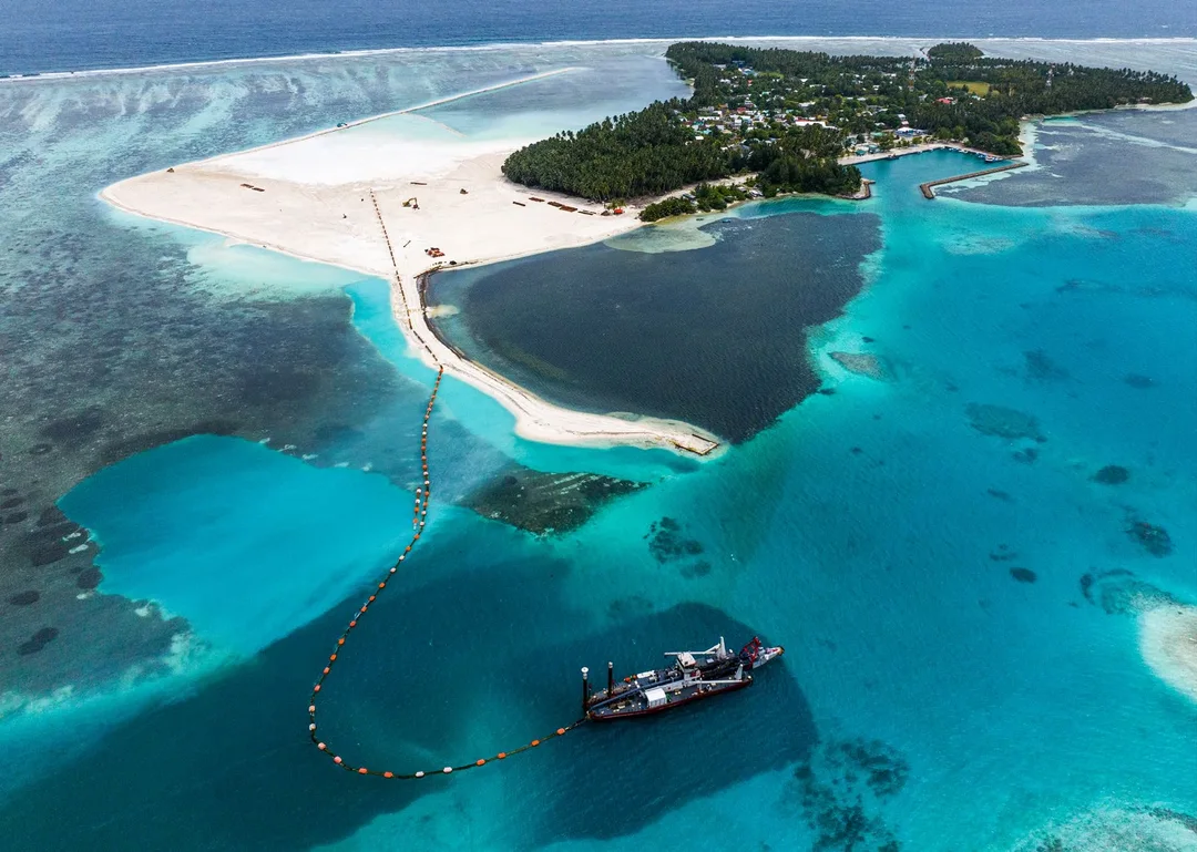 Aerial view of a dredger in a lagoon pumping sand through a long floating pipeline onto newly reclaimed white sand beside a wooded island.
