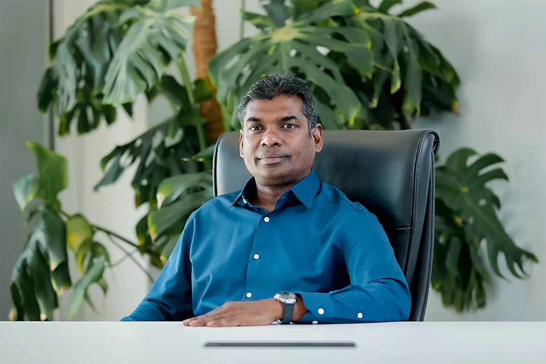A man in a blue shirt sits at a white desk in a black office chair, with large potted plants behind him.