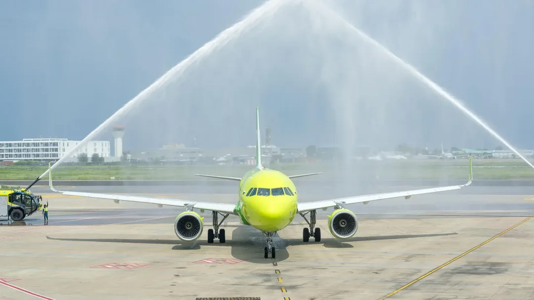 A lime-green S7 Airlines Airbus A320 taxis towards the camera under an arch of water sprayed by an airport fire truck, with a control tower and low airport buildings behind.