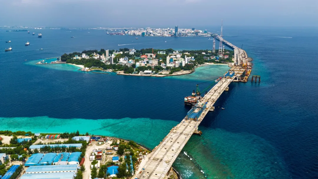Aerial view of a bridge deck running from an island with blue-roofed warehouses across the sea to a small, densely built island, with the tower blocks of Malé on the horizon.