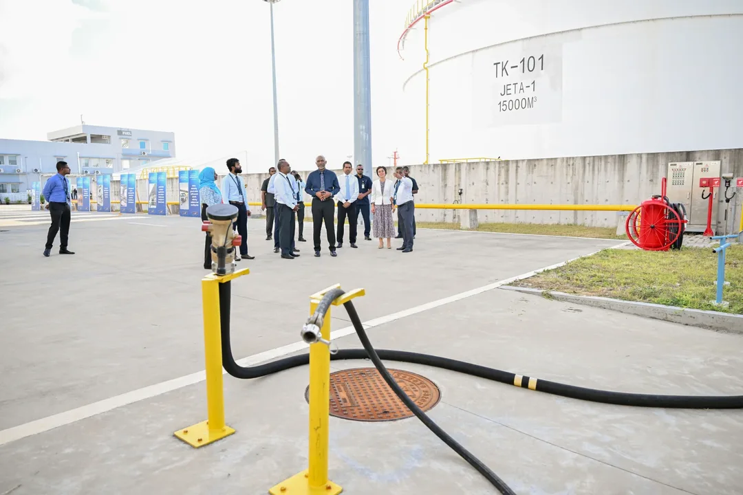 A group of officials stands in front of a large white storage tank marked "TK-101 JET A-1 15000M3", with a black fuel hose and yellow bollards in the foreground.